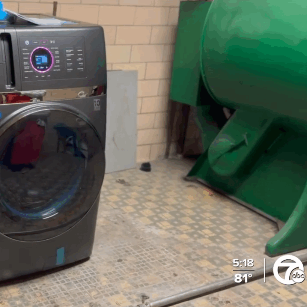 A modern front-loading washing machine is set against a tiled floor in what appears to be a laundry room. A large green industrial machine is visible in the background. The time and temperature are displayed in the bottom-right corner (5:18, 81 degrees). This setup could support the Detroit Public Schools' Laundry Initiative.