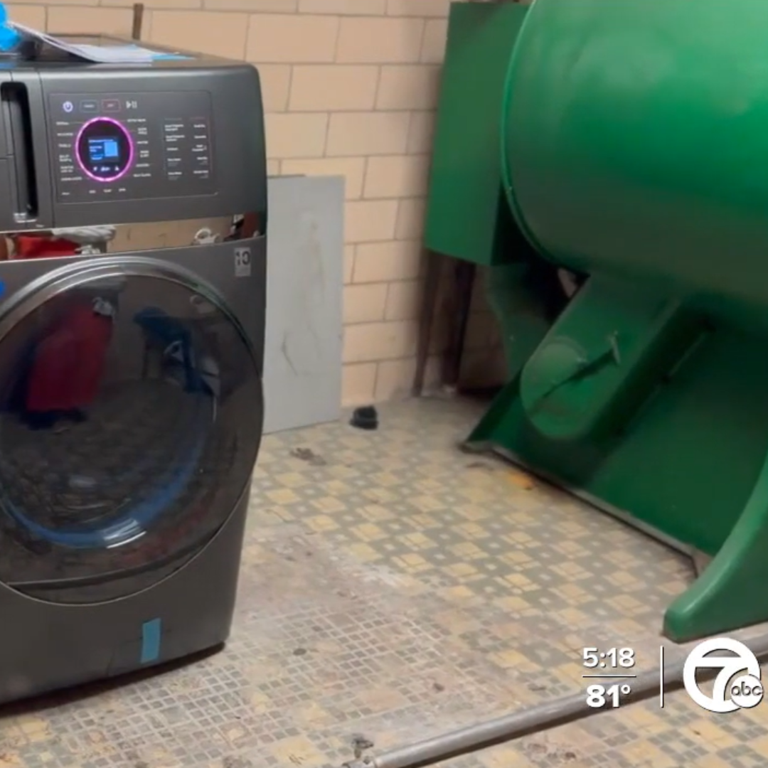 A modern front-loading washing machine is set against a tiled floor in what appears to be a laundry room. A large green industrial machine is visible in the background. The time and temperature are displayed in the bottom-right corner (5:18, 81 degrees). This setup could support the Detroit Public Schools' Laundry Initiative.