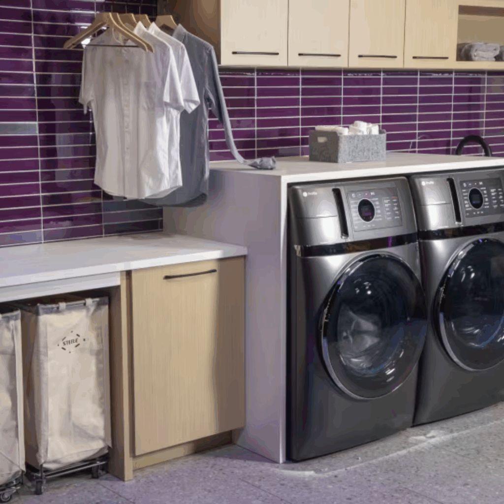 A modern laundry room with purple tiled backsplash, light wood cabinets, a white countertop, and new laundry machines. There are shirts hanging on a rod, laundry baskets below the counter, and a tissue box on top of the black front-loading washer and dryer.