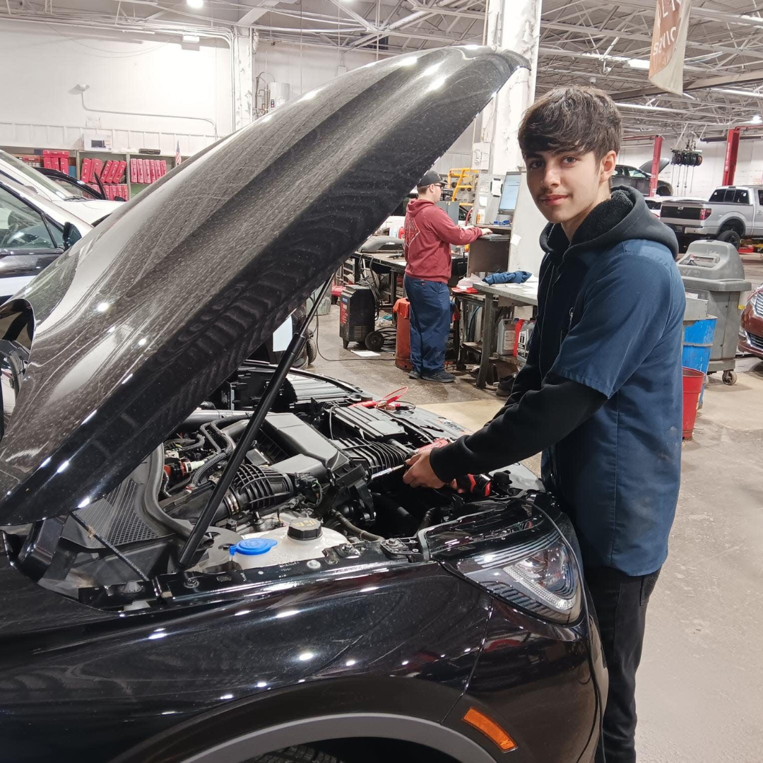 In an auto training center, a person in a blue mechanic jacket works diligently on a car with an open hood. Another individual observes in the background, while various tools and equipment fill the spacious garage, embodying the hands-on experience promoted by initiatives like the Joel Landy Foundation.