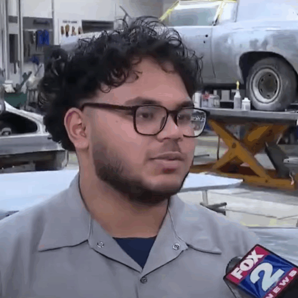 A person with curly hair and glasses speaks passionately in a workshop, preparing students for the automotive industry. In the background, a car glints under the spotlight while a "FOX 2 News" microphone captures every word.