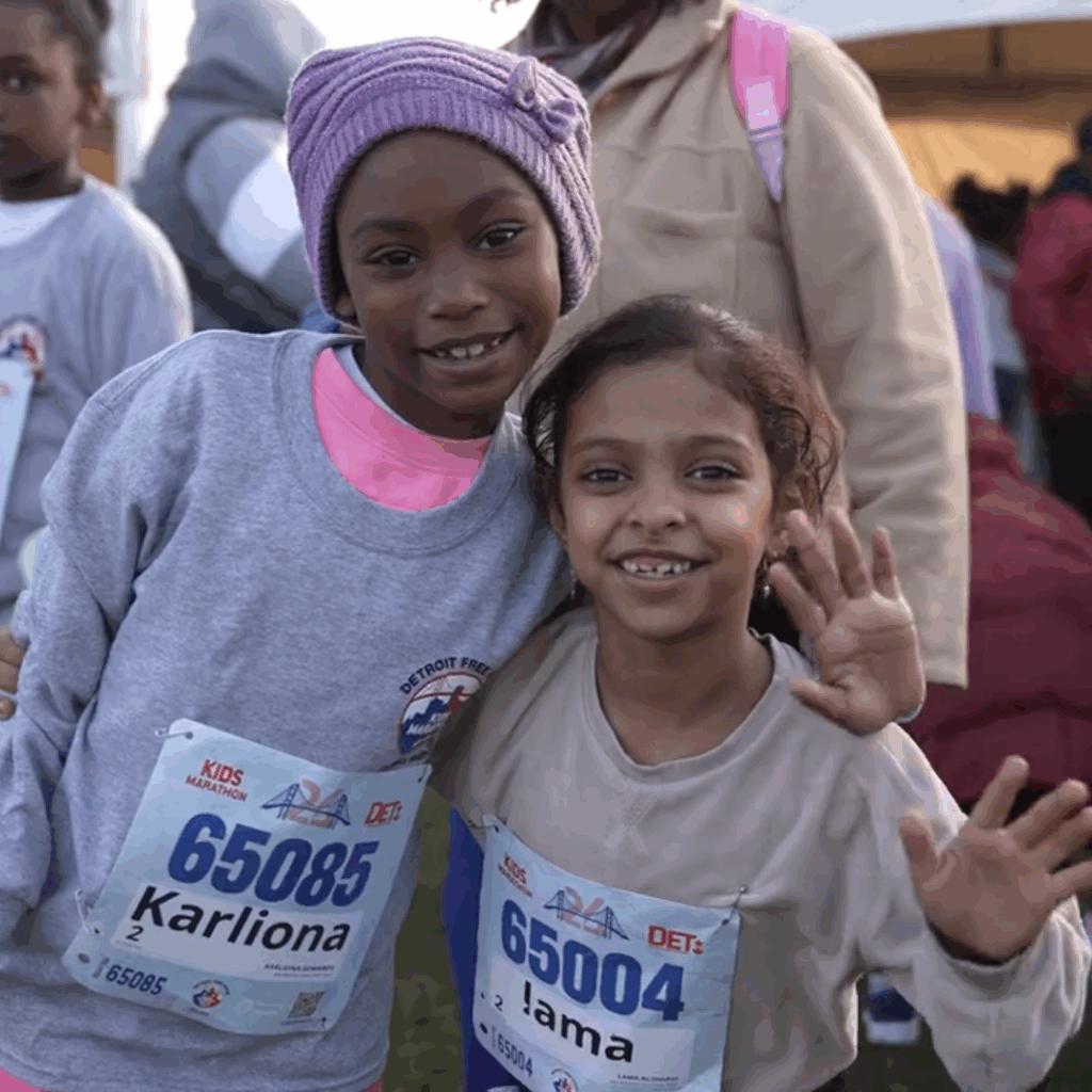 Two children wearing race bibs smile at the camera, embodying the spirit of the Detroit Free Press Marathon. The child on the left sports a purple hat and gray sweatshirt, while their friend in a beige shirt waves proudly. Their camaraderie reflects the joy of empowering Detroit's youth through running.