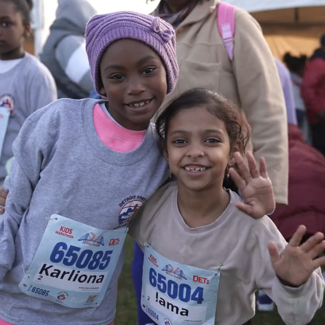 Two children wearing race bibs smile at the camera, embodying the spirit of the Detroit Free Press Marathon. The child on the left sports a purple hat and gray sweatshirt, while their friend in a beige shirt waves proudly. Their camaraderie reflects the joy of empowering Detroit's youth through running.