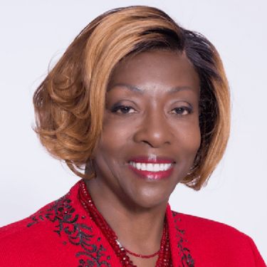 Smiling woman with wavy, shoulder-length light brown hair, wearing a red jacket with black embroidery and a red beaded necklace, against a plain white background—Dr. Iris Taylor, a dedicated nonprofit board leader.