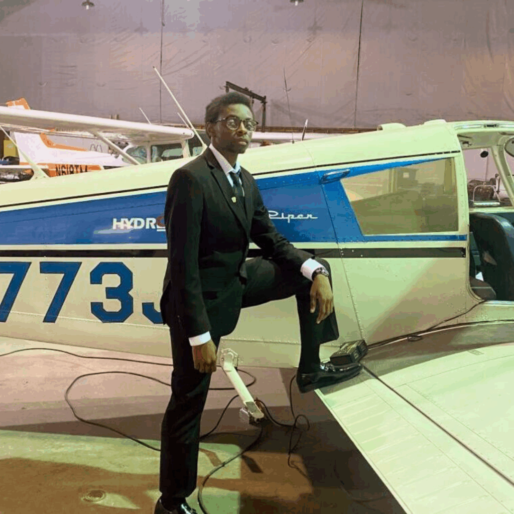 A man in a black suit poses confidently with one foot on the wing of a small white and blue airplane inside a hangar at Davis Aerospace, with other aircraft visible in the background.