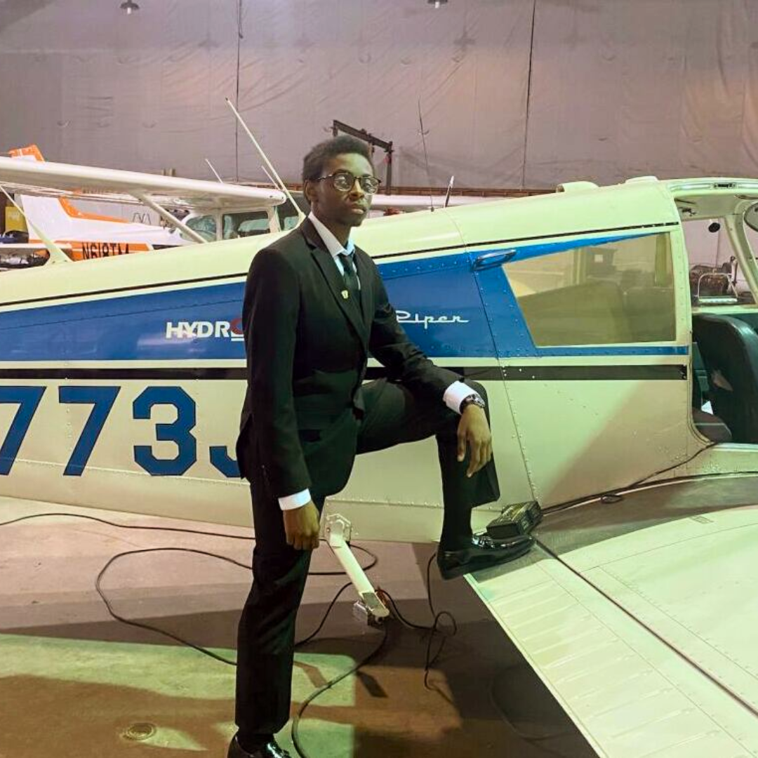 A man in a black suit poses confidently with one foot on the wing of a small white and blue airplane inside a hangar at Davis Aerospace, with other aircraft visible in the background.