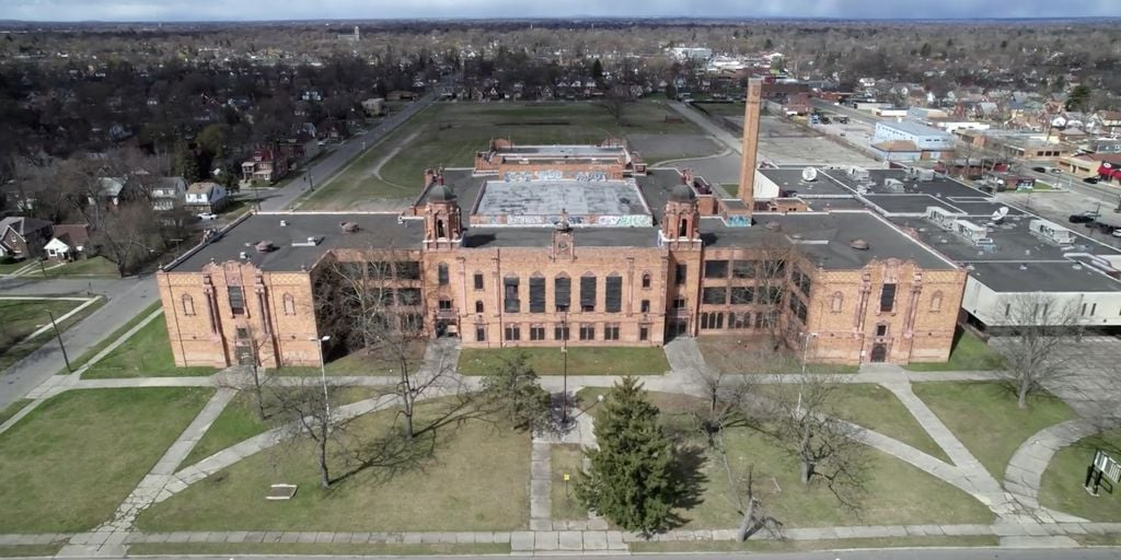 Aerial view of Cooley High School, a large, historic red-brick building with a tall smokestack, surrounded by leafless trees, grassy areas, and intersecting sidewalks in a suburban Detroit Schools neighborhood.