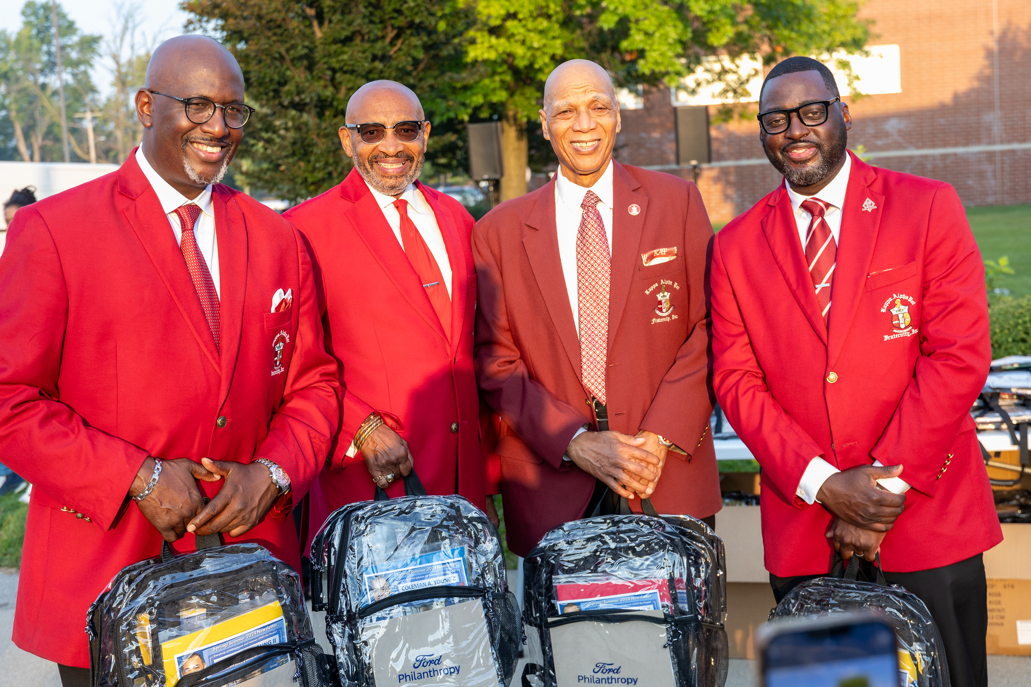 Four men in red blazers standing together outside a Detroit public school with student backpacks