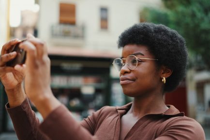 Beautiful dark haired girl in glasses taking photo for her blog outdoors. Afro woman using smartphone on city street
